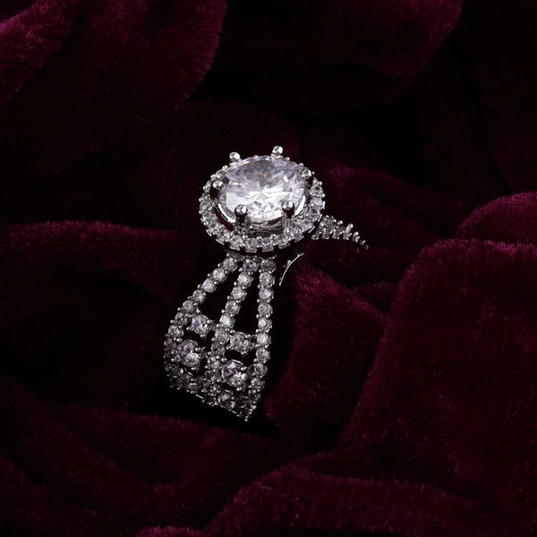 Elegant silver-toned statement ring featuring a large round sparkling center stone, surrounded by crystal embellishments and a detailed dangling design, displayed on dark velvet fabric.
